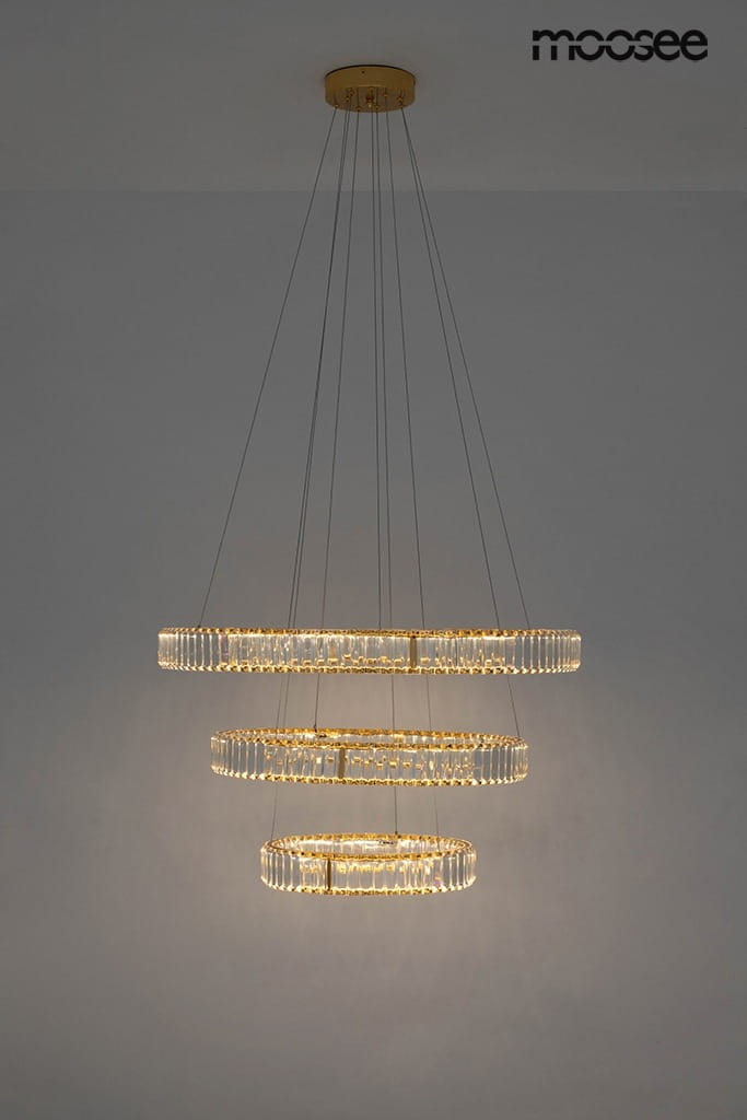 Lampa wisząca Liberty Triples LED z trzema okrągłymi pierścieniami, wykonana z przezroczystego szkła i złotego metalu. Zawieszona na cienkich linkach, emituje ciepłe światło, idealna do nowoczesnych wnętrz.