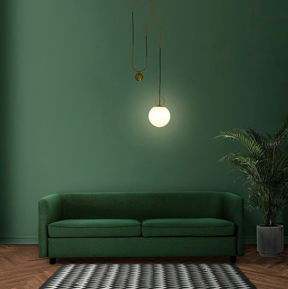Lampa wisząca Lift 1-punktowa w kolorze złotym z okrągłym, białym kloszem. Zawieszona na cienkim przewodzie w minimalistycznym stylu. Tło w odcieniu zieleni, obok ciemnozielona kanapa i roślina w doniczce.