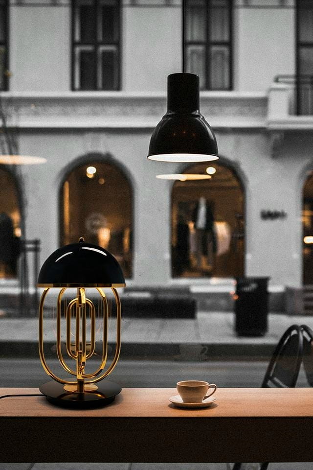 Lampa stołowa Bottega 2-punktowa w kolorze złotym i czarnym, z okrągłą podstawą i elegancką, zaokrągloną kloszem. Ustawiona na drewnianym blacie obok filiżanki kawy, w tle widoczne okna z delikatnym oświetleniem.
