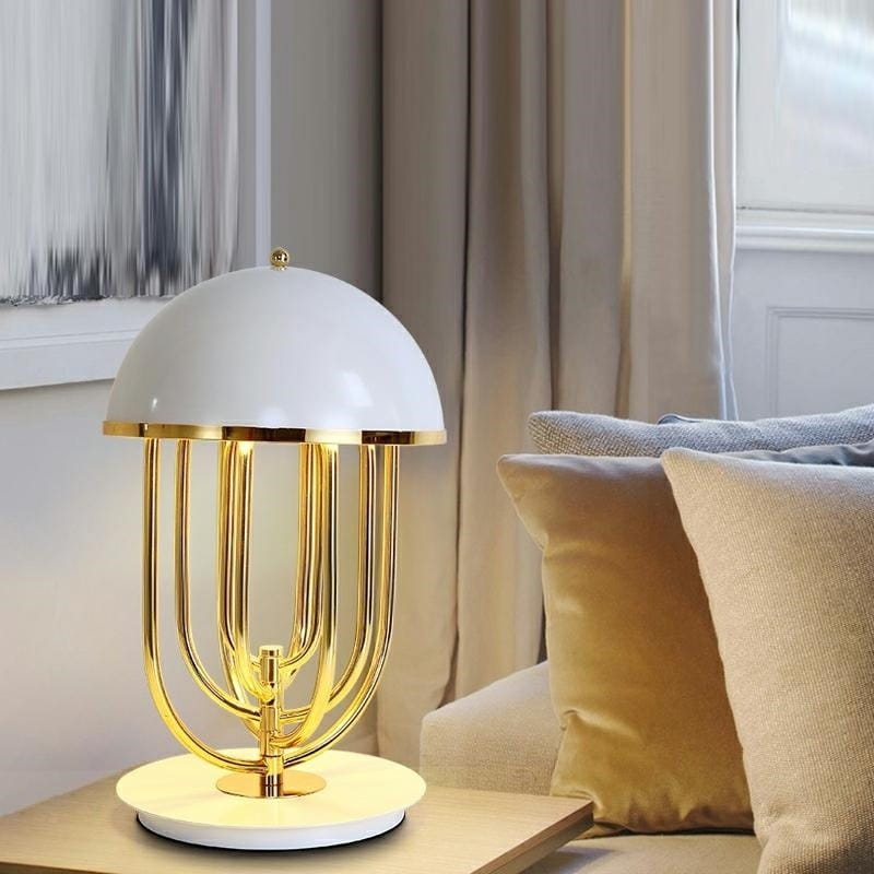 Lampa stołowa Bottega 2-punktowa w kolorze złotym i białym, z zaokrąglonym kloszem i metalową podstawą. Wykonana z błyszczącego metalu, z widocznymi złotymi elementami. Ustawiona na stoliku obok miękkiej kanapy, w tle jasne zasłony.
