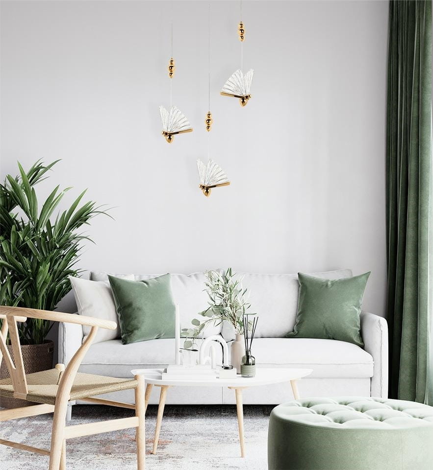 Złota lampa wisząca Butterfly M LED z białymi kloszami w kształcie skrzydeł motyla. Zawieszona w nowoczesnym wnętrzu, nad sofą, wśród zielonych roślin i akcesoriów. Prosta, elegancka forma w minimalistycznym stylu.