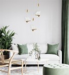 Złota lampa wisząca Butterfly M LED z białymi kloszami w kształcie skrzydeł motyla. Zawieszona w nowoczesnym wnętrzu, nad sofą, wśród zielonych roślin i akcesoriów. Prosta, elegancka forma w minimalistycznym stylu.