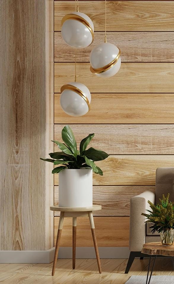 Lampa wisząca Globe 20 L w złotym kolorze, z trzema okrągłymi kloszami w białym odcieniu, zawieszona nad donicą z rośliną. Klosze mają złote zdobienia. Lampa znajduje się w nowoczesnym wnętrzu z drewnianą ścianą i meblami.