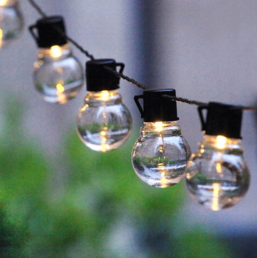 Girlanda ogrodowa solarna z 20 kulkami LED na czarnym przewodzie, długość 4,85 m. Kuleczki w przezroczystym, okrągłym kształcie z czarnymi uchwytami, zawieszone na delikatnym sznureczku. Tło z rozmytymi zielonymi roślinami.