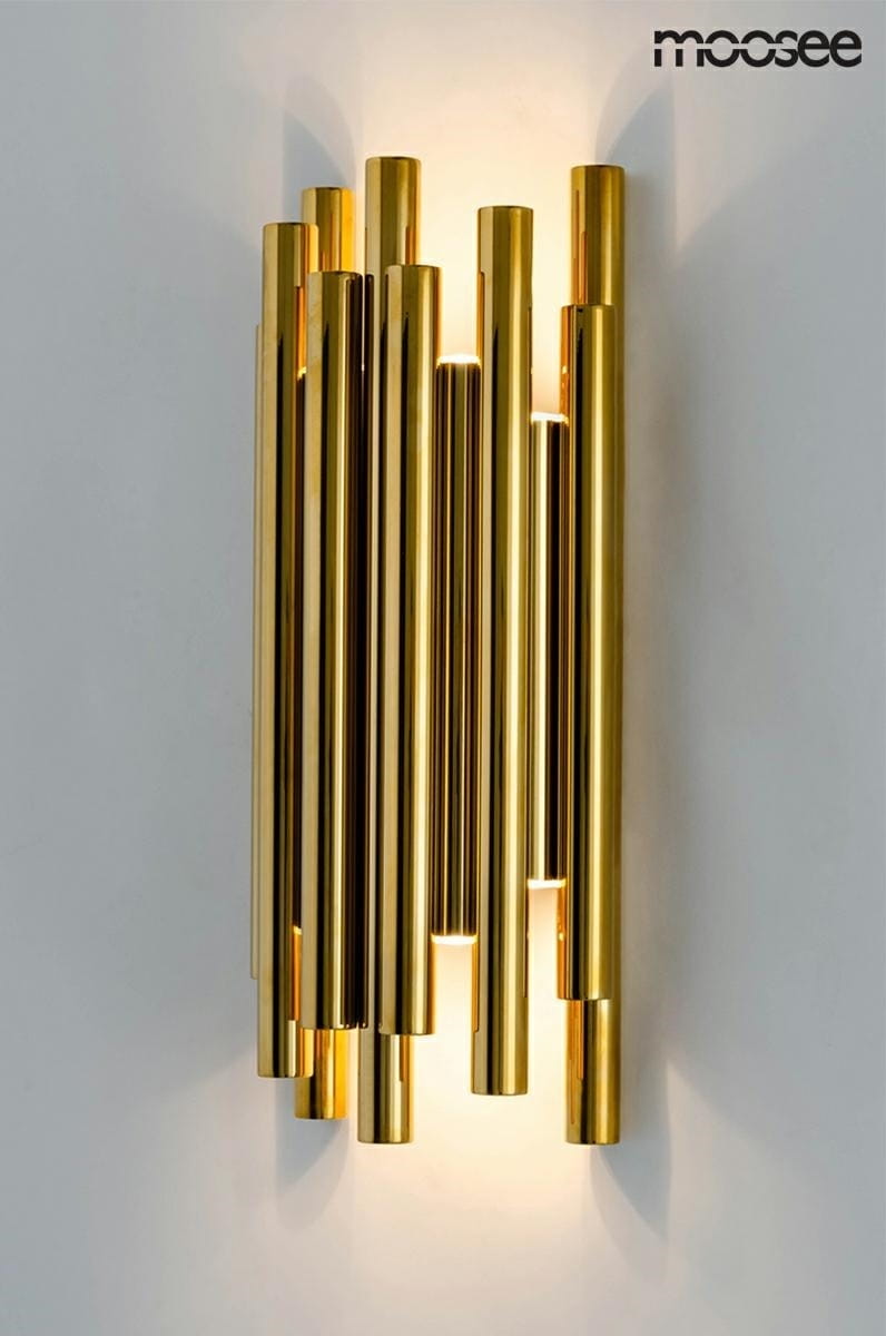 Kinkiet LED Organo 8-punktowy w kolorze złotym, o cylindrycznym kształcie, z ośmioma wykończeniami w różnych długościach. Zamontowany na białej ścianie, emituje ciepłe światło z tyłu, tworząc efektowne oświetlenie.