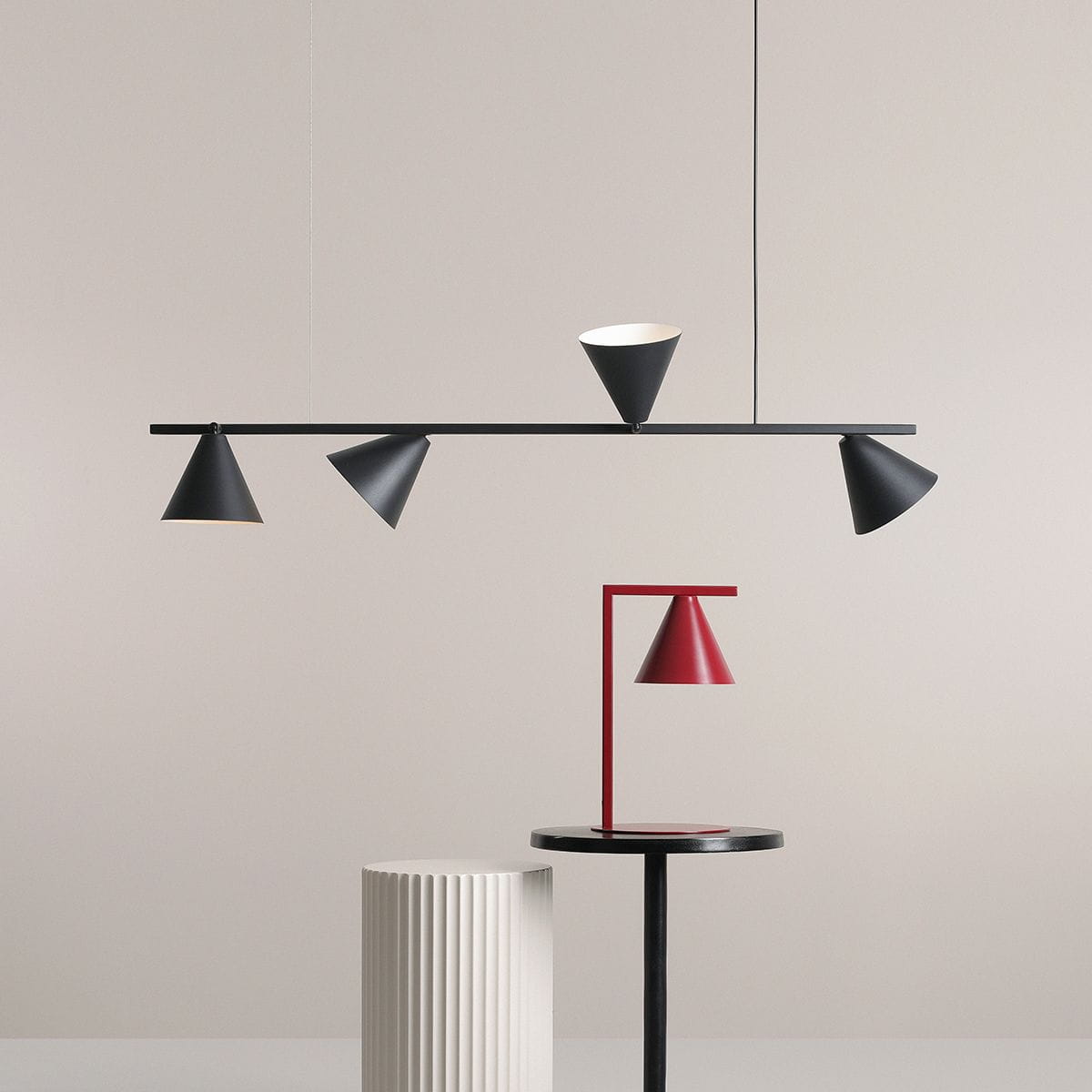 Czarna lampa wisząca 4-punktowa Form Black 1108L1 z minimalistycznymi, stożkowymi kloszami w czerni i bieli zawieszona na cienkim stelażu. W tle widoczna czerwona lampa stołowa na czarnej podstawie, na jasnym tle.