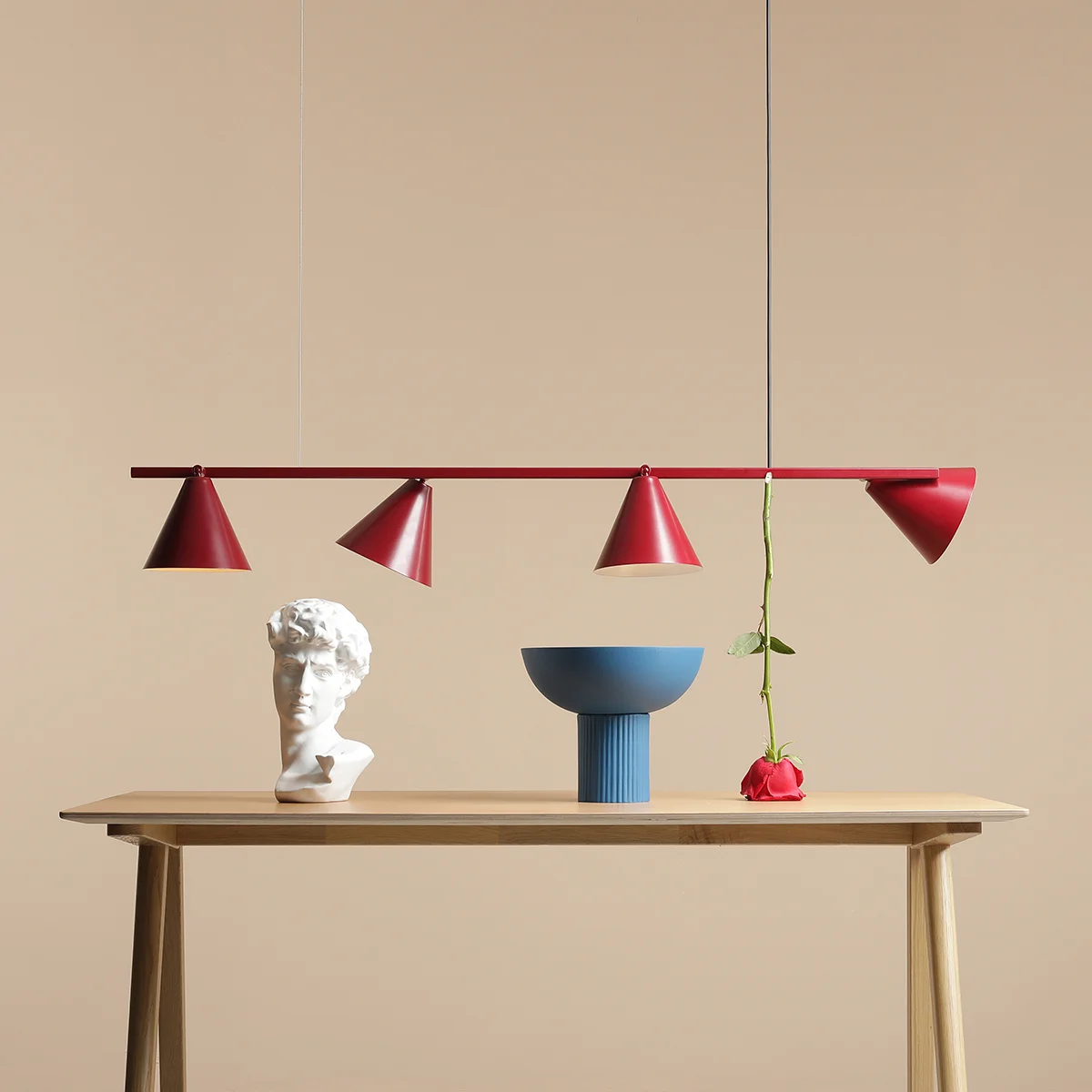 Lampa wisząca Form Red Wine, 4-punktowa, z czterema czerwonymi kloszami w kształcie stożków zawieszona na cienkiej belce. W tle widoczna drewniana ława z białą rzeźbą, niebieską misą i różą. Tło w odcieniu beżowym.