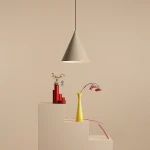 Lampa wisząca Form Beige 1-punktowa, w kształcie stożka, w kolorze beżowym. Zawieszona na cienkim przewodzie nad minimalistycznym schodkiem z dekoracyjnymi wazonami w kolorach czerwonym i żółtym. Tło w odcieniu beżowym.