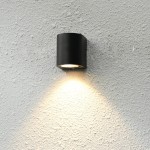 Kinkiet elewacyjny Genta w kolorze czarnym, okrągły, zamontowany na szarej ścianie. Emituje światło skierowane w dół, tworząc efektowny cień na powierzchni. Wysokiej jakości materiał, minimalistyczny design, idealny do użytku zewnętrznego.