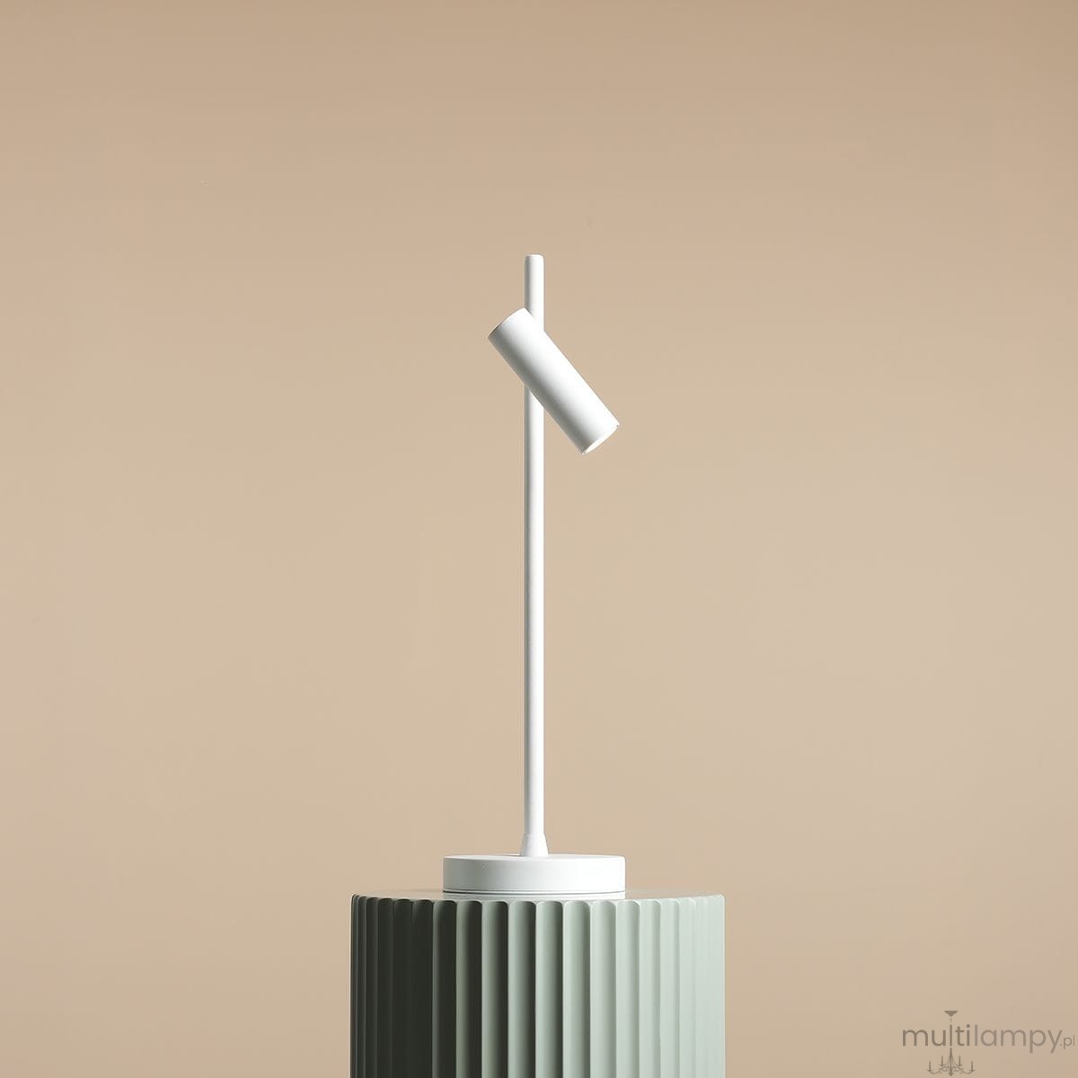 Lampka biurkowa Trevo 1104B w kolorze białym, o cylindrycznym kloszu zamontowanym pod kątem na prostym, smukłym stojaku. Ustawiona na zielonym, teksturowanym podłożu, na jasnym tle.