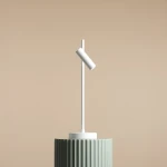 Lampka biurkowa Trevo 1104B w kolorze białym, o cylindrycznym kloszu zamontowanym pod kątem na prostym, smukłym stojaku. Ustawiona na zielonym, teksturowanym podłożu, na jasnym tle.