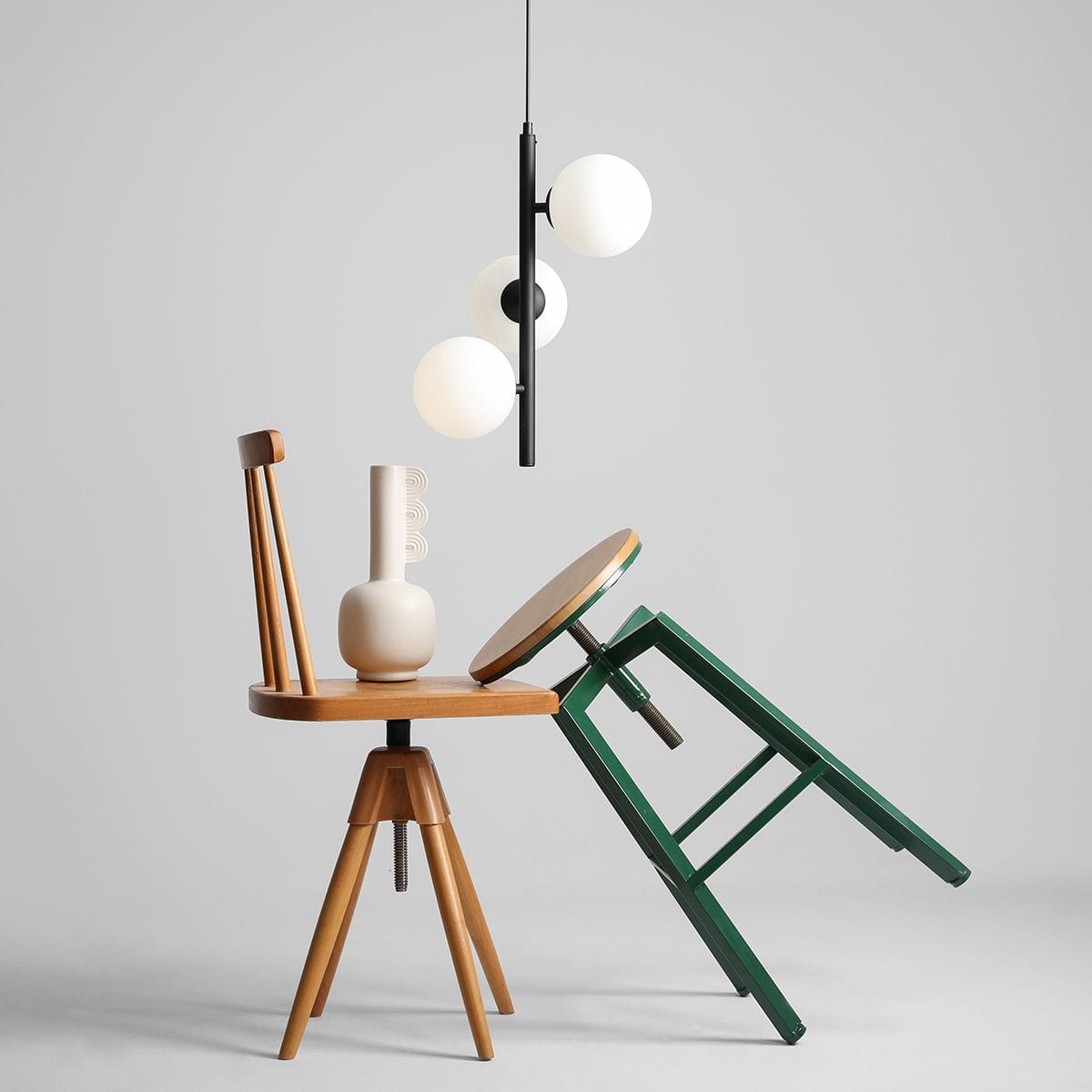 Lampa wisząca Fin S 3-punktowa w kolorze czarnym z trzema okrągłymi kloszami w odcieniu białym. Zawieszona pod kątem nad drewnianymi krzesłami w różnych pozycjach, na szarym tle.
