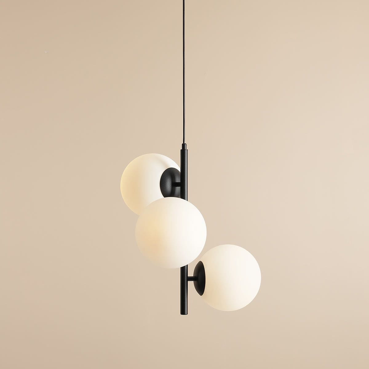 Lampa wisząca Fin M, 3-punktowa, w kolorze czarnym. Składa się z trzech białych, szklanych kul zamocowanych na czarnym, metalowym stelażu. Ujęcie z przodu, wisząca na cienkim kablu, na tle w neutralnym beżu.