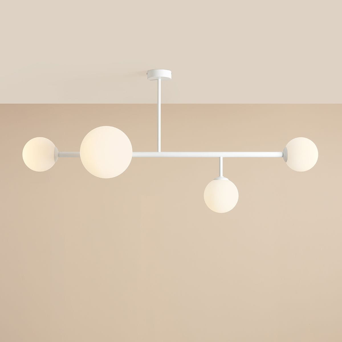 Lampa sufitowa Dione 4-punktowa w kolorze białym z okrągłymi kloszami. Wyposażona w cztery źródła światła umieszczone na prostokątnej konstrukcji, montowana sufitowo. Klosze w matowym wykończeniu, rozłożone na różnych wysokościach. Tło w jasnym odcieniu.