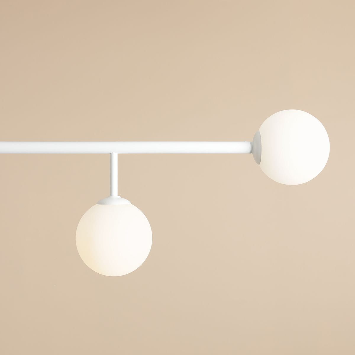 Lampa sufitowa Dione 4-punktowa w kolorze białym. Posiada dwa okrągłe klosze z matowego szkła zawieszone na prostym, białym stelażu. Ujęcie z góry, ukazujące detale konstrukcji i kształt kulistych kloszy. Tło w odcieniu beżu.