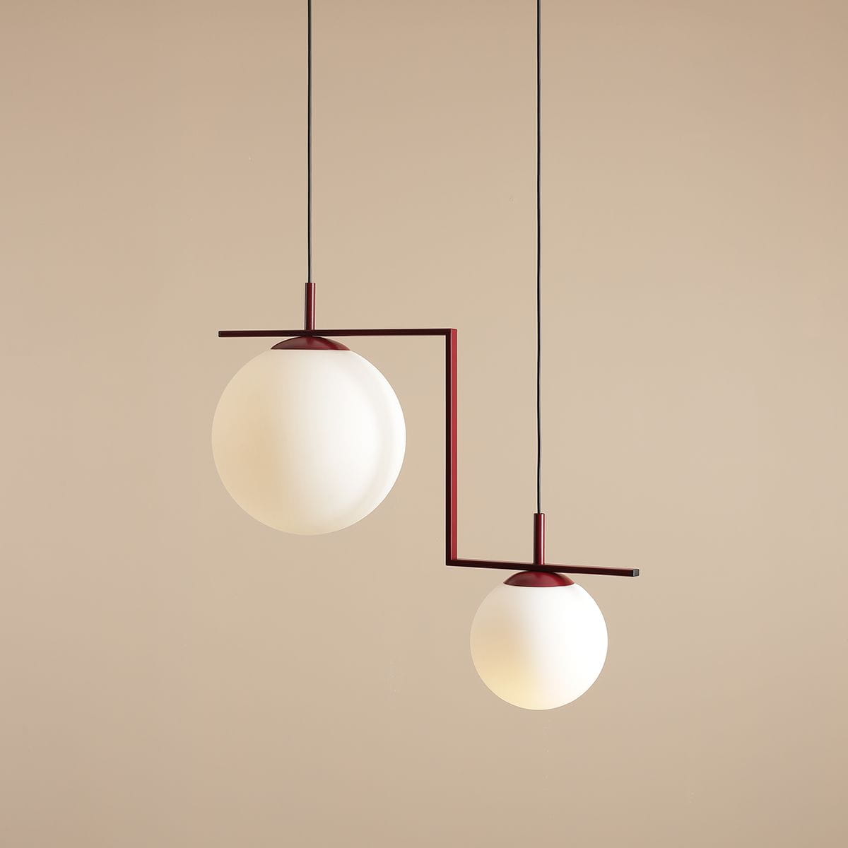 Lampa wisząca Zac II 2-punktowa w kolorze czerwonym, z dwiema białymi kulami szklanymi. Zawieszona na cienkich czarnych przewodach, z geometrycznym metalowym stelażem. Górna kula większa, dolna mniejsza, układ asymetryczny. Tło jasne, jednolite.