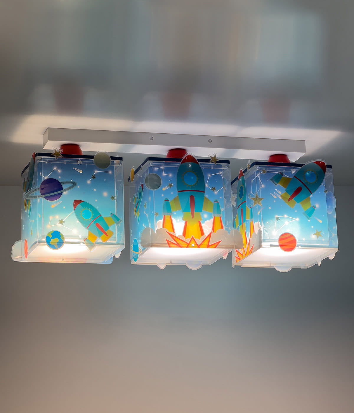 Lampa sufitowa 3-punktowa z motywem rakiet i planet. Kwadratowe klosze w błękitnym kolorze z grafiką rakiet, gwiazd i planet. Ułożona na białej konstrukcji, z lampami w środku, emitującymi ciepłe światło.