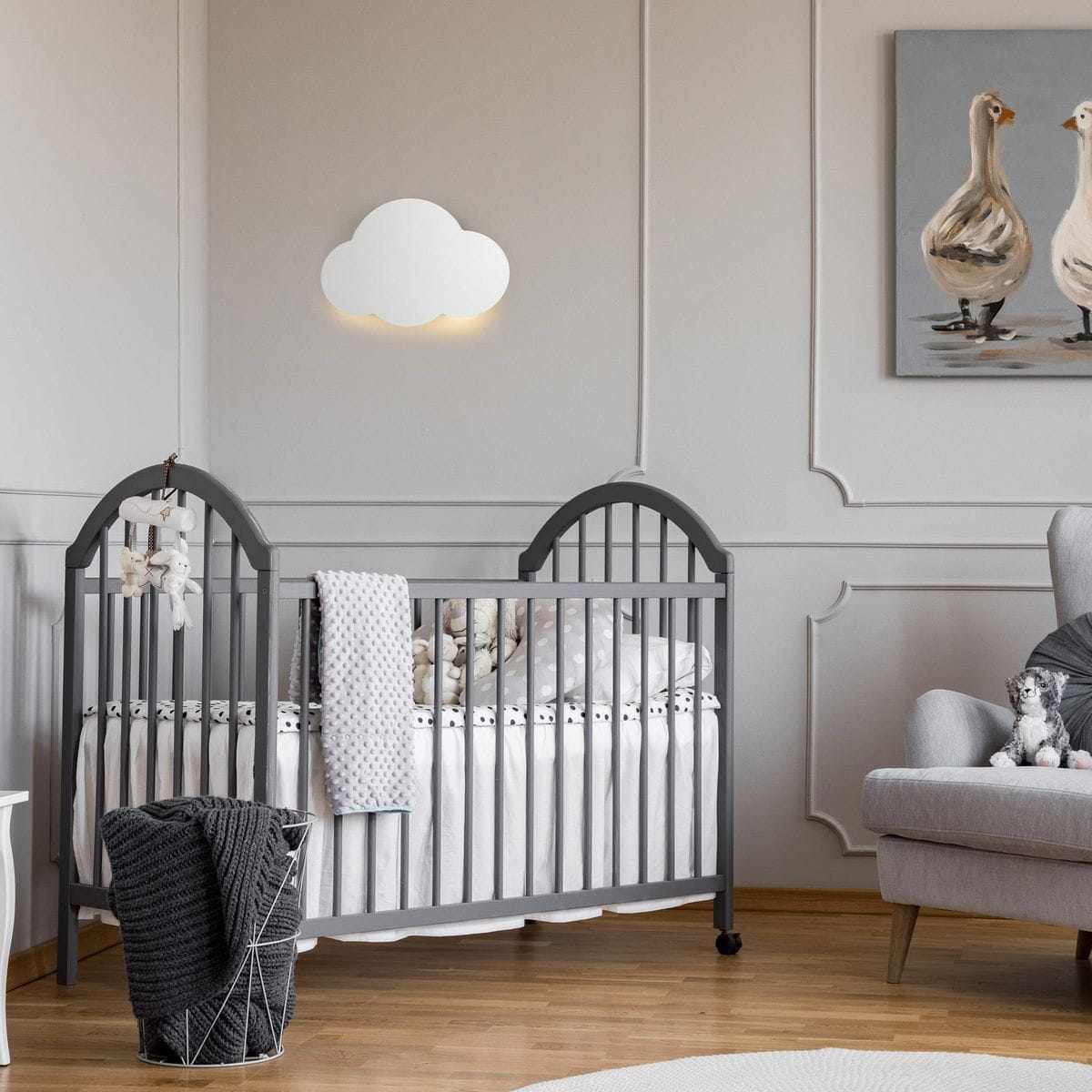 Biały kinkiet w kształcie chmurki, montowany na ścianie, emitujący delikatne światło. Znajduje się nad łóżeczkiem dziecięcym w jasnym, minimalistycznym wnętrzu. Tło w neutralnych kolorach, z elementami dekoracyjnymi.