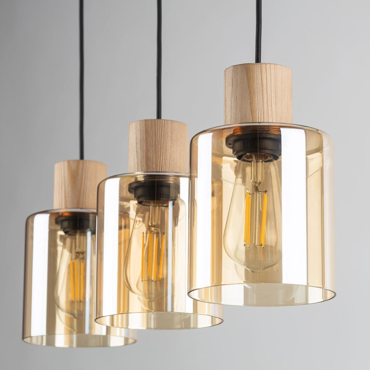 Lampa wisząca Orson 3-punktowa w kolorze czarnym i bursztynowym. Szklane klosze w odcieniu bursztynu z drewnianymi elementami, zawieszone na czarnych przewodach. Klosze mają cylindryczny kształt, eksponując żarówki. Tło jasne, minimalistyczne.
