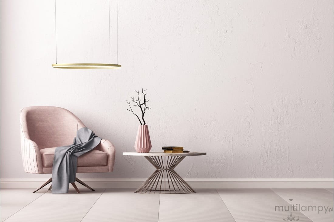 Złota lampa wisząca Halo D80 LED, umieszczona nad stołem, w minimalistycznym wnętrzu. Lampa ma okrągły kształt, zawieszona na cienkich linkach. W tle widoczny różowy fotel z szarym kocem oraz różowa wazon z gałązką. Ściana ma jasny, gładki kolor.
