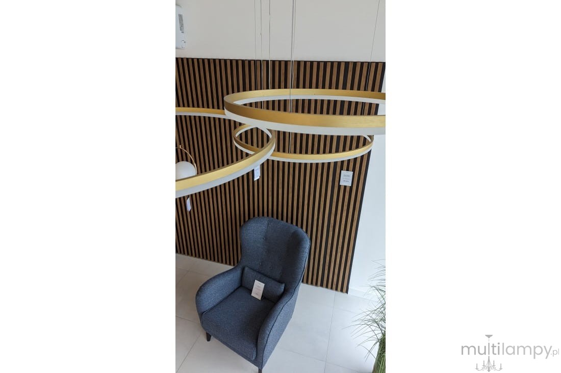 Lampa wisząca Halo 100+80+60 LED w kolorze złotym, składająca się z trzech okrągłych pierścieni, zawieszona na cienkich linkach. Ujęcie z góry, w tle widoczny niebieski fotel na tle drewnianej, poziomej ścianki.