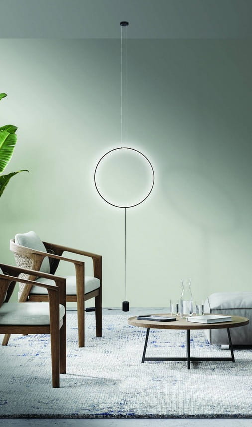 Czarna lampa wisząca Codutti LED 1-punktowa, w kształcie okręgu, zawieszona na cienkim przewodzie. W tle nowoczesny salon z drewnianymi krzesłami i okrągłym stolikiem na jasnym dywanie. Ściana w delikatnym odcieniu zieleni.