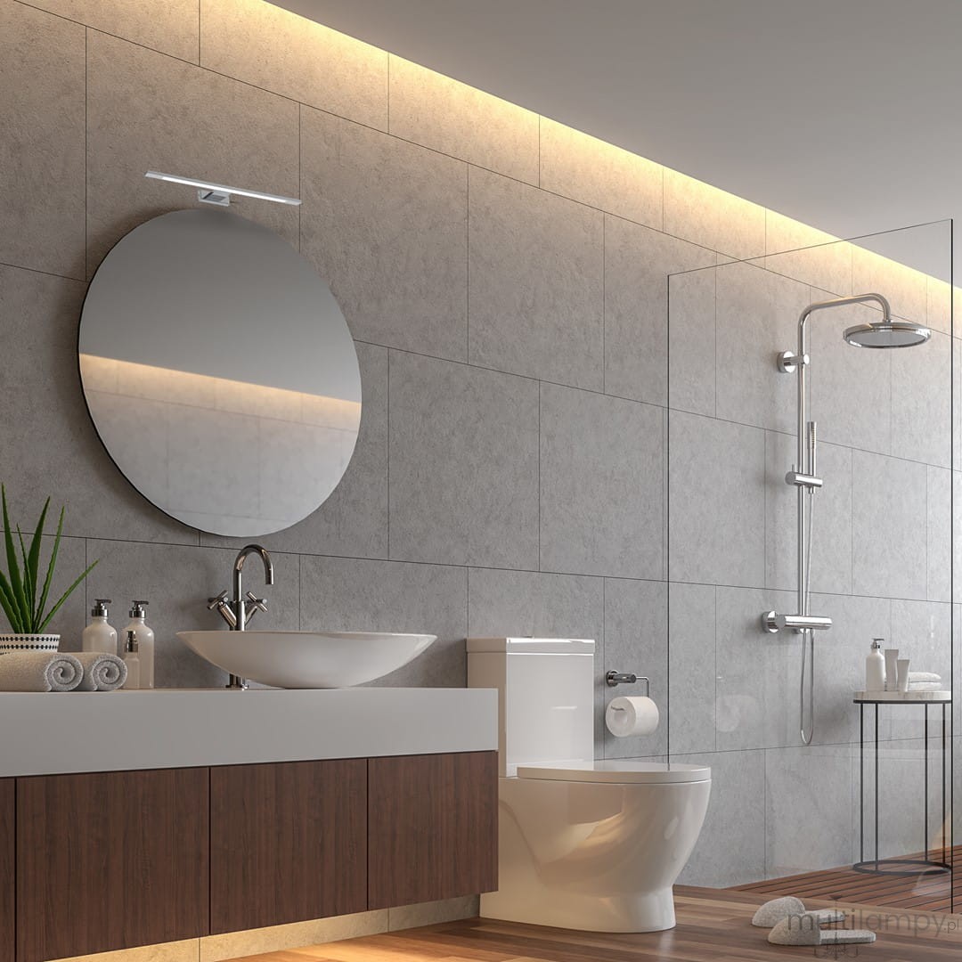 Kinkiet Shine LED IP44 w kolorze chromu, zamontowany nad okrągłym lustrem w nowoczesnej łazience. Produkt ma prosty, smukły kształt i emituje jasne światło. Tło stanowi szara płytka ścienna, a w aranżacji widoczna umywalka i toaleta.