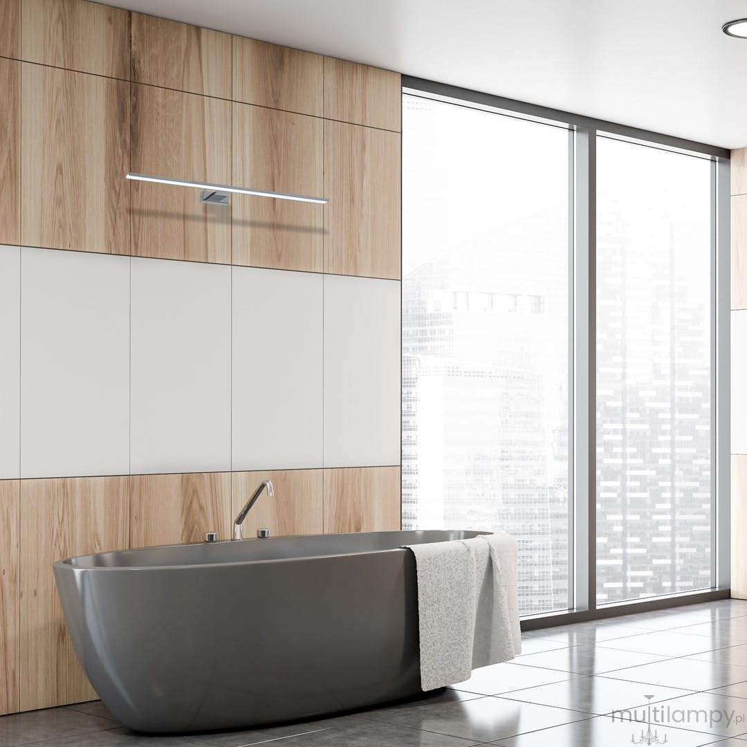 Kinkiet Shine LED IP44 w chromowym wykończeniu, montowany na ścianie w łazience. Długi, prostokątny kształt, umieszczony nad wanną. W tle drewniane panele i duże okno z widokiem na miasto.