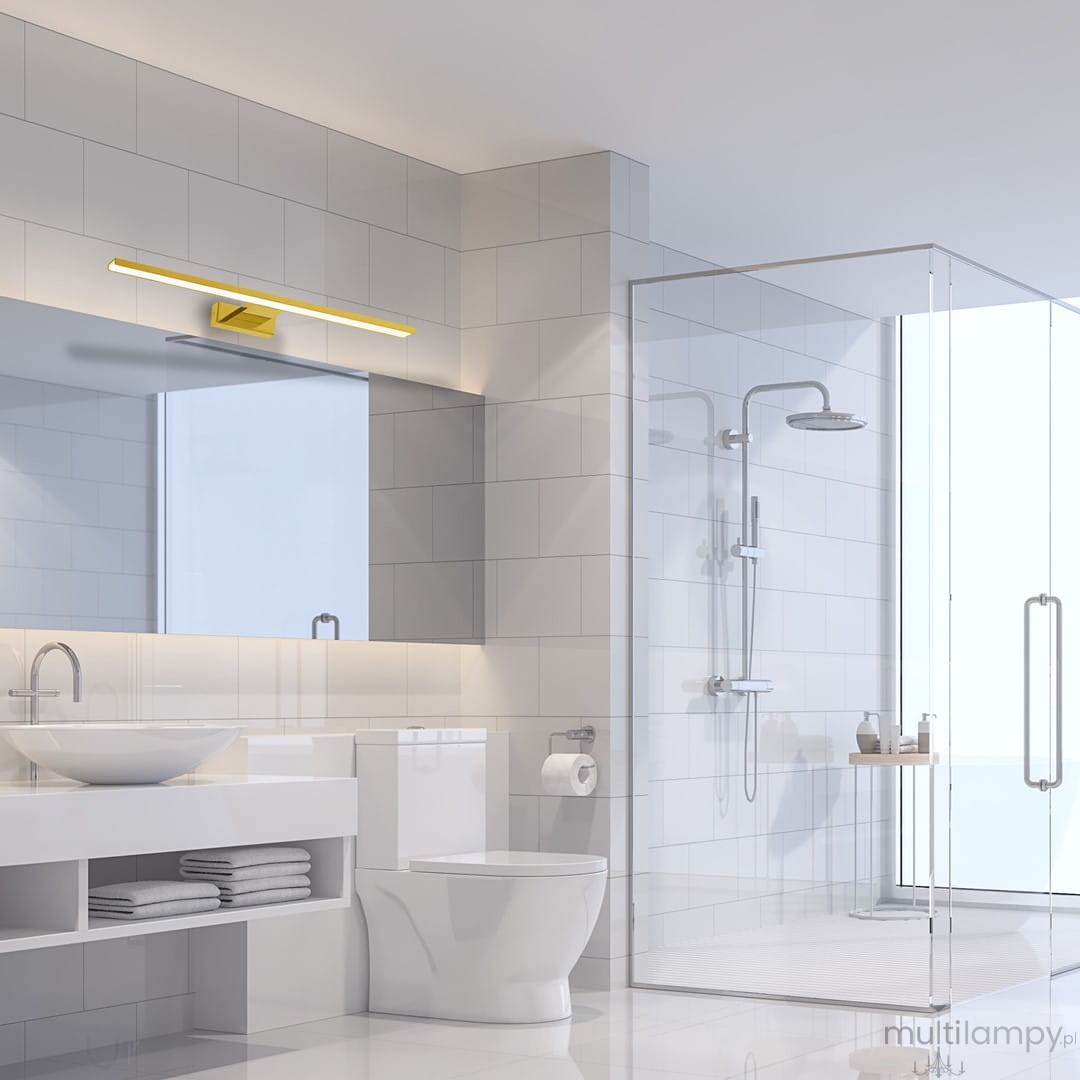 Kinkiet Shine LED IP44 w kolorze złotym, montowany na ścianie, z prostokątnym kloszem. Zainstalowany nad lustrem w nowoczesnej łazience, na jasnym tle płytek. Oświetlenie skierowane w dół, podkreślające elegancki design.