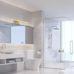 Kinkiet Shine LED IP44 w kolorze złotym, montowany na ścianie, z prostokątnym kloszem. Zainstalowany nad lustrem w nowoczesnej łazience, na jasnym tle płytek. Oświetlenie skierowane w dół, podkreślające elegancki design.