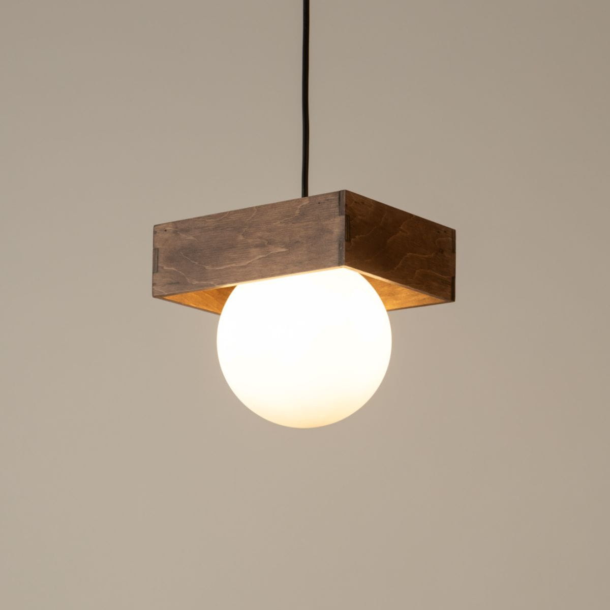 Czarna/brązowa lampa wisząca Bloom z okrągłym kloszem z białego szkła. Ujęcie z dołu, produkt zawieszony na czarnym przewodzie. Kwadratowa drewniana podstawa z widocznym usłojeniem, nadająca rustykalny charakter. Tło w jasnym odcieniu.