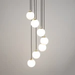 Czarna lampa wisząca Piano z siedmioma okrągłymi kloszami w białym kolorze, zawieszona na cienkich, czarnych przewodach. Klosze mają złote detale u góry, tworząc nowoczesny wygląd. Lampa jest zawieszona w pionie, na jednolitym, jasnoszarym tle.