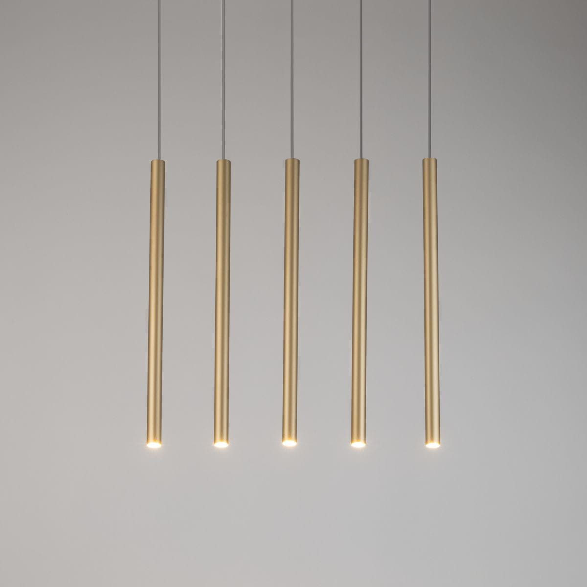 Złota lampa wisząca Sopel Prosty, pięciopunktowa, złożona z pięciu smukłych cylindrów zawieszonych na cienkich linkach. Kąt ujęcia frontalny, na jednolitym tle. Widoczne źródła światła na dole każdego cylindra.