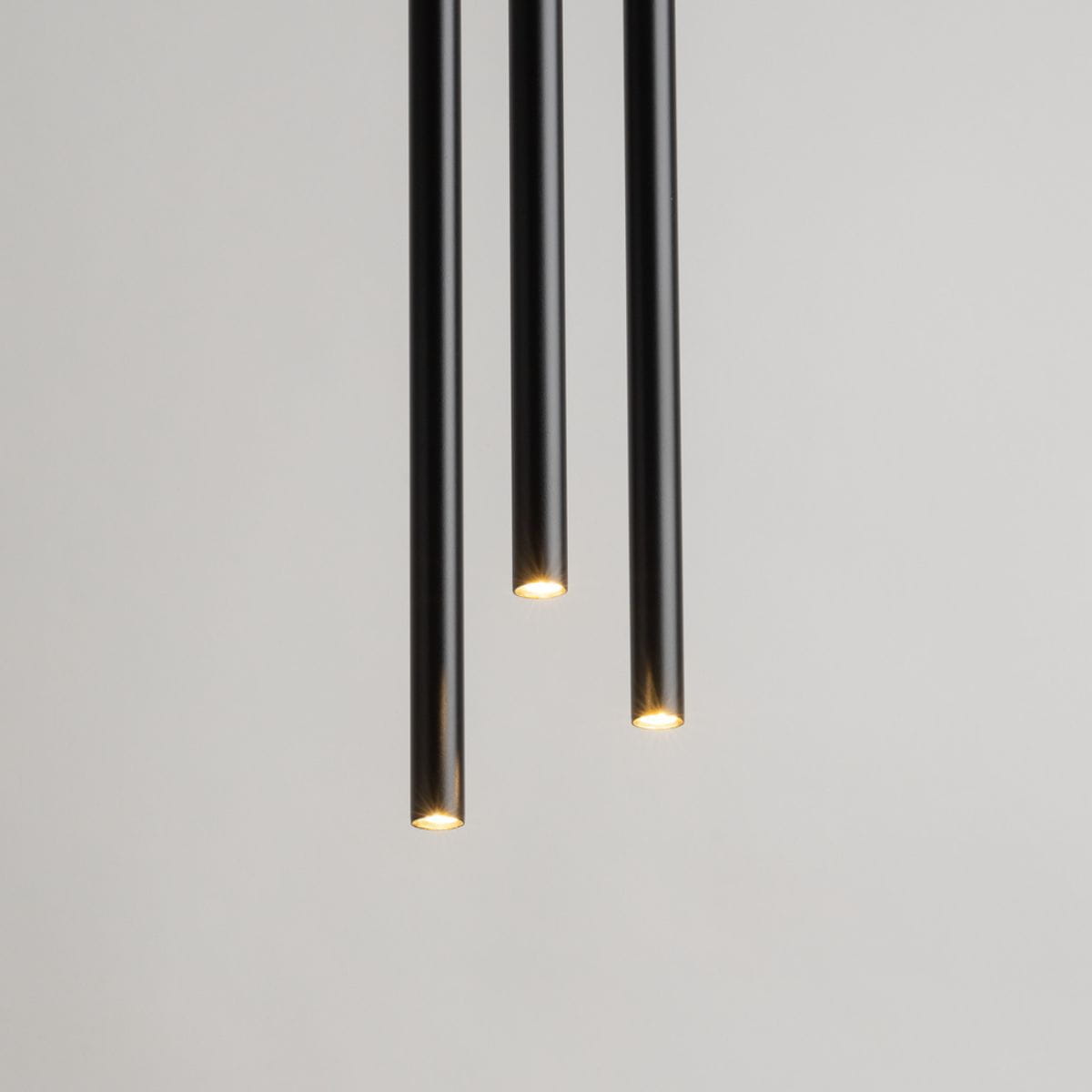 Czarna, wisząca lampa 3-punktowa Sopel Koło z trzema cylindrycznymi kloszami, emitującymi ciepłe światło. Lampa zawieszona w pionie, z minimalistycznym designem, na jasnym tle.