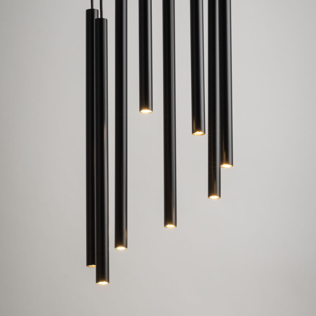 Lampa wisząca Sopel Koło, 8-punktowa, czarna. Składa się z ośmiu cylindrycznych elementów w różnych długościach, emitujących światło z końców. Zawieszona na cienkich linkach, w minimalistycznym stylu, na jasnym tle.