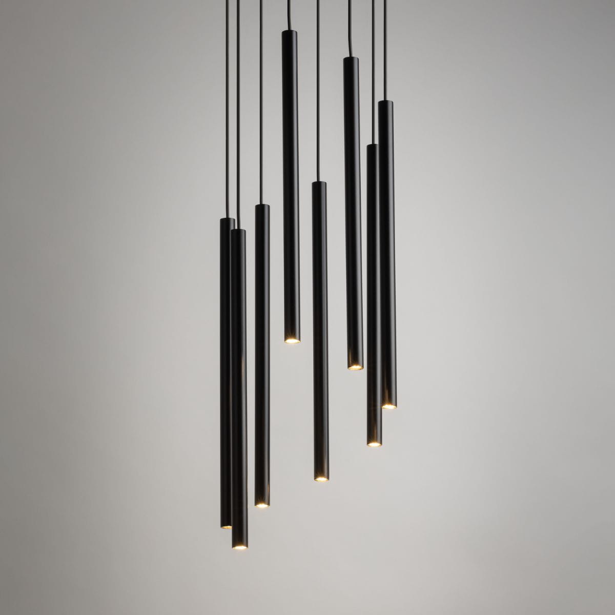 Lampa wisząca Sopel Koło, 8-punktowa, czarna. Składa się z ośmiu długich, cylindrycznych źródeł światła, zawieszonych na cienkich linkach. Całość ma nowoczesny design, z delikatnym podświetleniem na końcach. Tło jasne, minimalistyczne.