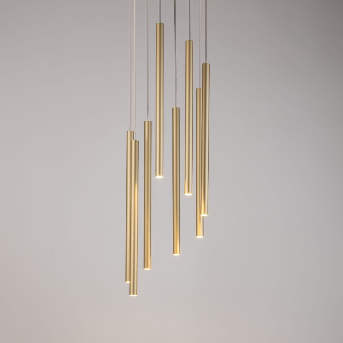 Lampa wisząca Sopel Koło z ośmioma złotymi, cylindrycznymi kloszami, zwisająca na cienkich linkach. Klosze mają różne długości i emitują światło z dolnej krawędzi, na tle jasnej ściany.