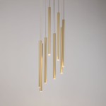 Lampa wisząca Sopel Koło z ośmioma złotymi, cylindrycznymi kloszami, zwisająca na cienkich linkach. Klosze mają różne długości i emitują światło z dolnej krawędzi, na tle jasnej ściany.