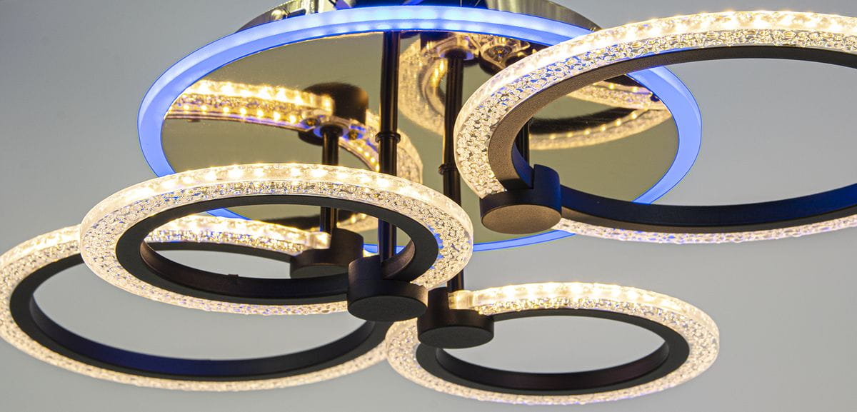 Lampa sufitowa z pilotem, 4 okrągłe pierścienie LED w kolorze złotym i czarnym. Okręgi mają dekoracyjne, przezroczyste wstawki oraz niebieskie podświetlenie. Ujęcie z dołu, ukazujące szczegóły konstrukcji i źródła światła.