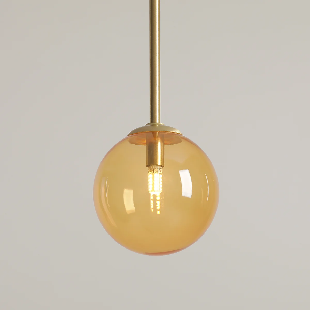 Lampa wisząca Bosso D14 z mosiężnym zawieszeniem i szklanym kloszem w kolorze pomarańczowym. Okrągła forma z widoczną żarówką G9, zawieszona na długim stelażu. Tło jasne, jednolite.