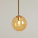 Lampa wisząca Bosso D14 z mosiężnym zawieszeniem i szklanym kloszem w kolorze pomarańczowym. Okrągła forma z widoczną żarówką G9, zawieszona na długim stelażu. Tło jasne, jednolite.