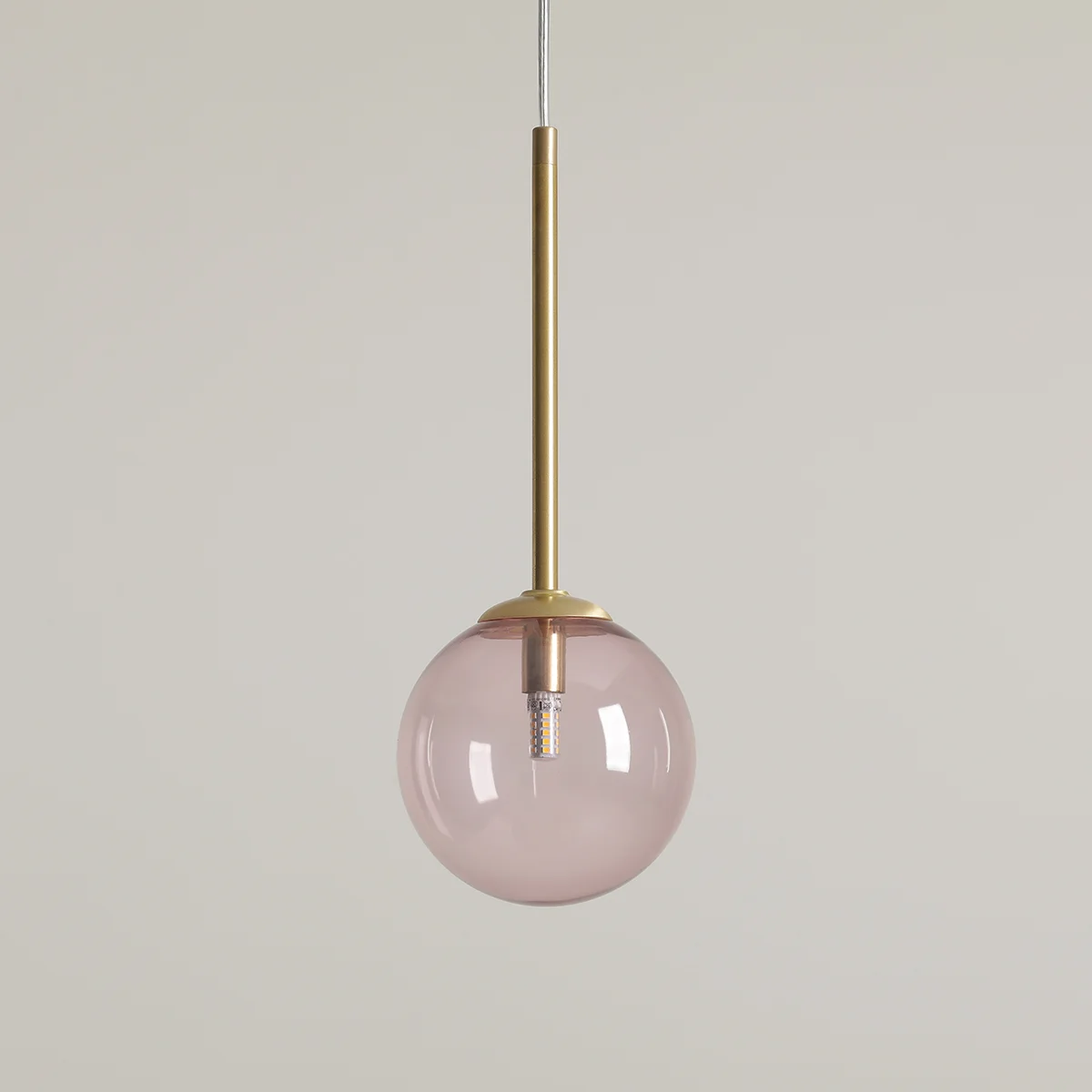 Lampa wisząca Bosso D14 w kolorze mosiądzu i różowym. Szklana kula z przezroczystym wnętrzem zawieszona na cienkim, metalowym słupku. Kąt ujęcia z przodu, produkt wisi na białym tle. Wnętrze lampy widoczne.