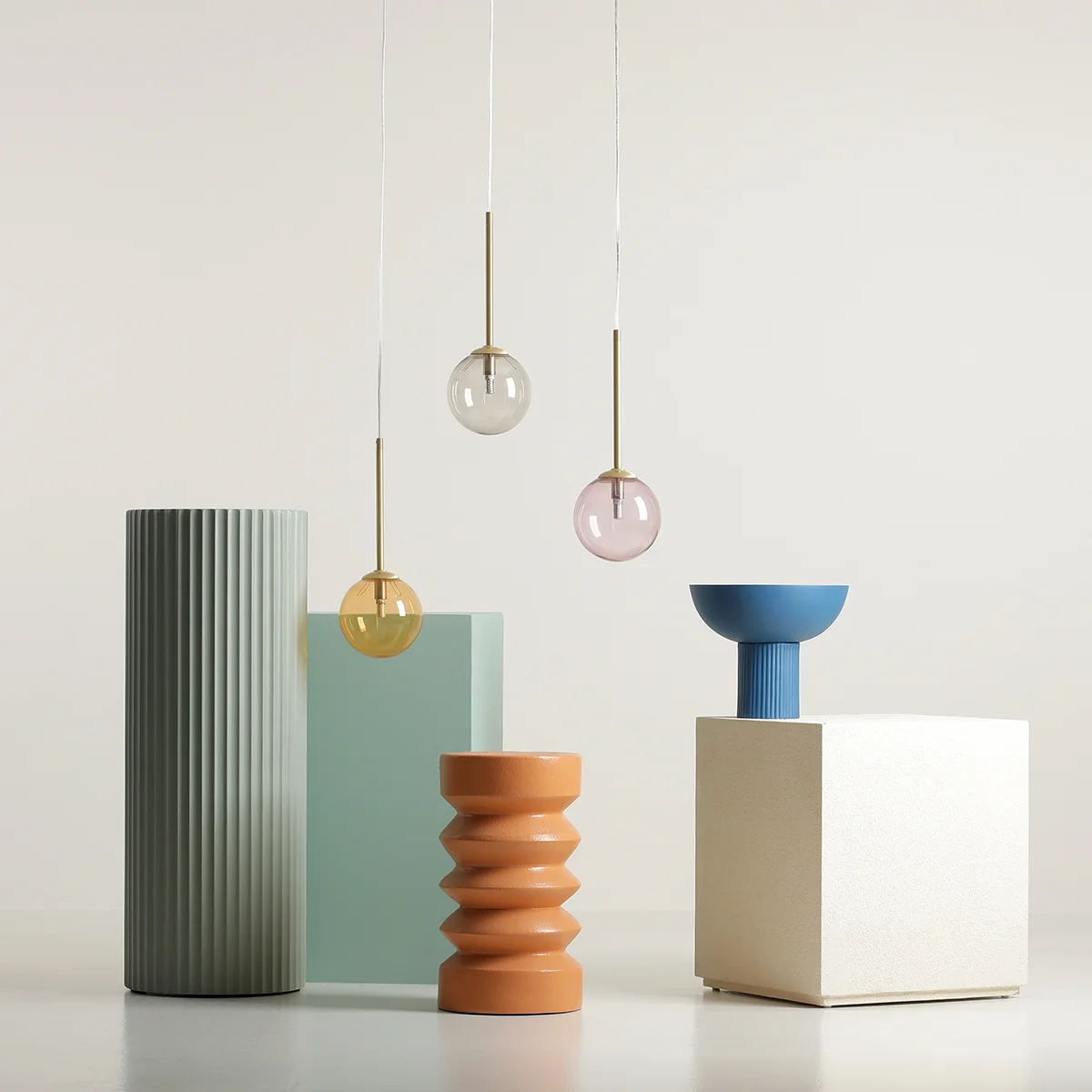 Lampa wisząca Bosso 3xG9 w kolorze mosiądzu z wielokolorowymi kloszami (żółty, różowy, przezroczysty) zawieszona nad różnorodnymi geometrycznymi podstawami w pastelowych odcieniach, na jasnym tle.