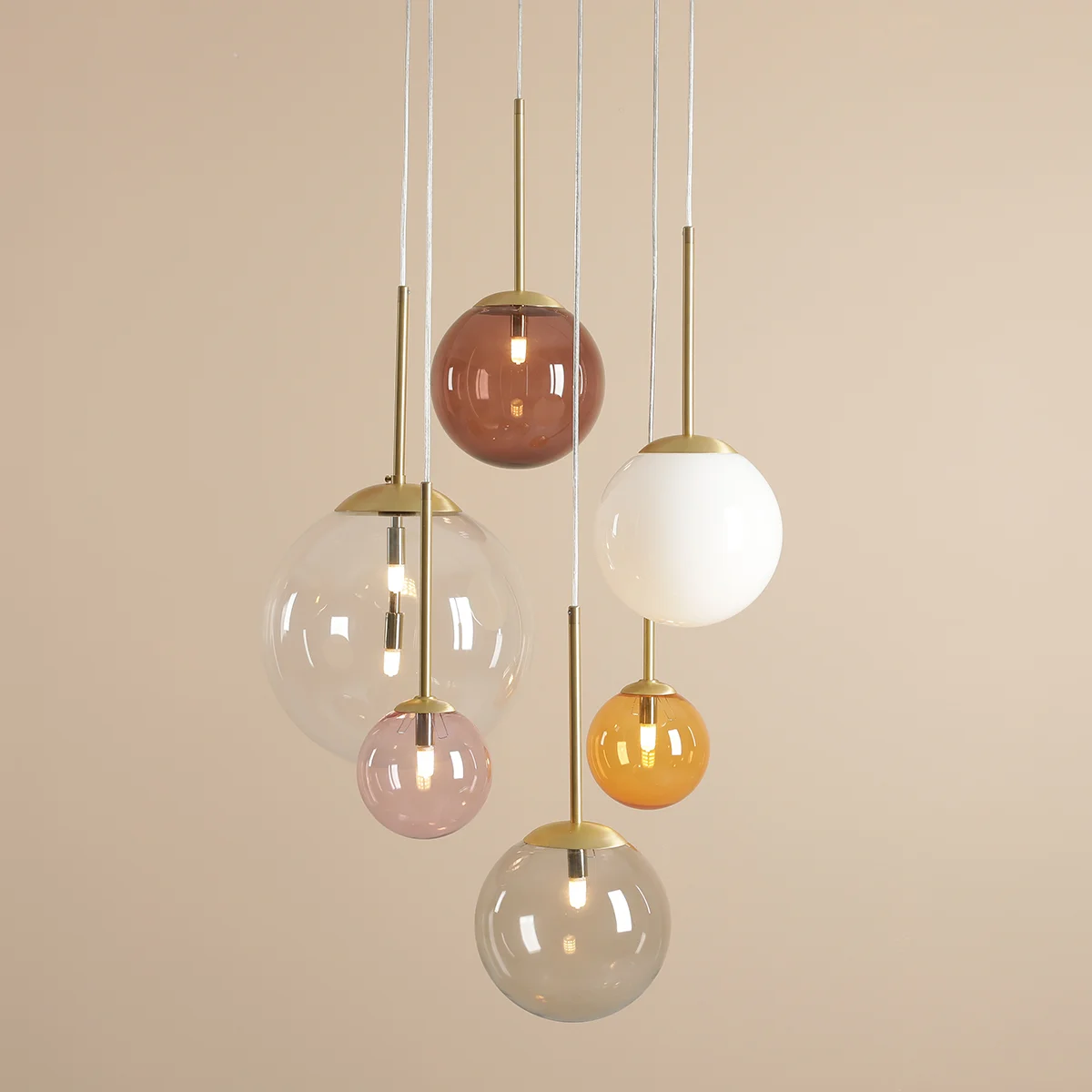Lampa wisząca Bosso w kolorze mosiądzu z pięcioma kulami G9 i jedną E27, zawieszona na białych linkach. Kule w różnych odcieniach, w tym przezroczysta, różowa, brązowa, biała i pomarańczowa, tworzą nowoczesny, multikolorowy efekt.