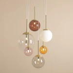Lampa wisząca Bosso w kolorze mosiądzu z pięcioma kulami G9 i jedną E27, zawieszona na białych linkach. Kule w różnych odcieniach, w tym przezroczysta, różowa, brązowa, biała i pomarańczowa, tworzą nowoczesny, multikolorowy efekt.