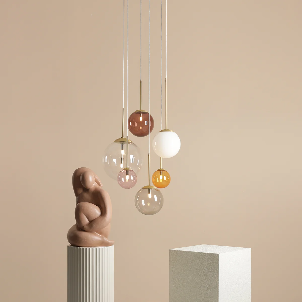 Lampa wisząca Bosso z pięcioma szklanymi kulami w różnych kolorach (różowy, brązowy, przezroczysty, biały, pomarańczowy) oraz złotymi elementami. Zawieszona na cienkich sznurach, w nowoczesnym otoczeniu z minimalistycznymi rzeźbami.