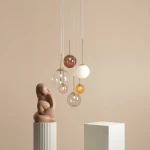 Lampa wisząca Bosso z pięcioma szklanymi kulami w różnych kolorach (różowy, brązowy, przezroczysty, biały, pomarańczowy) oraz złotymi elementami. Zawieszona na cienkich sznurach, w nowoczesnym otoczeniu z minimalistycznymi rzeźbami.