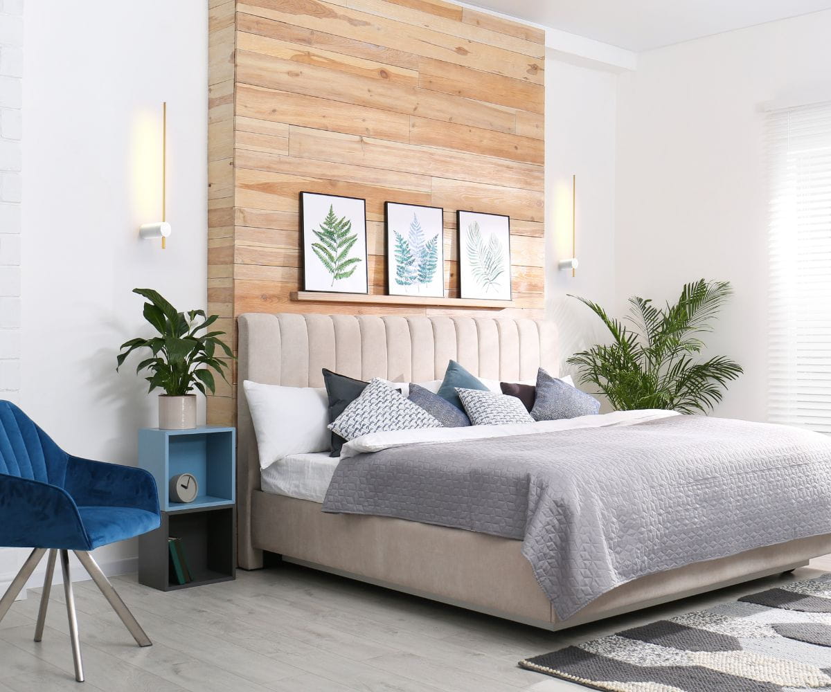 Kinkiet LED Wand 466 w kolorze białym i złotym, montowany na ścianie, z prostą, minimalistyczną formą. Widoczny w aranżacji sypialni obok łóżka i rośliny, na tle drewnianej ściany.