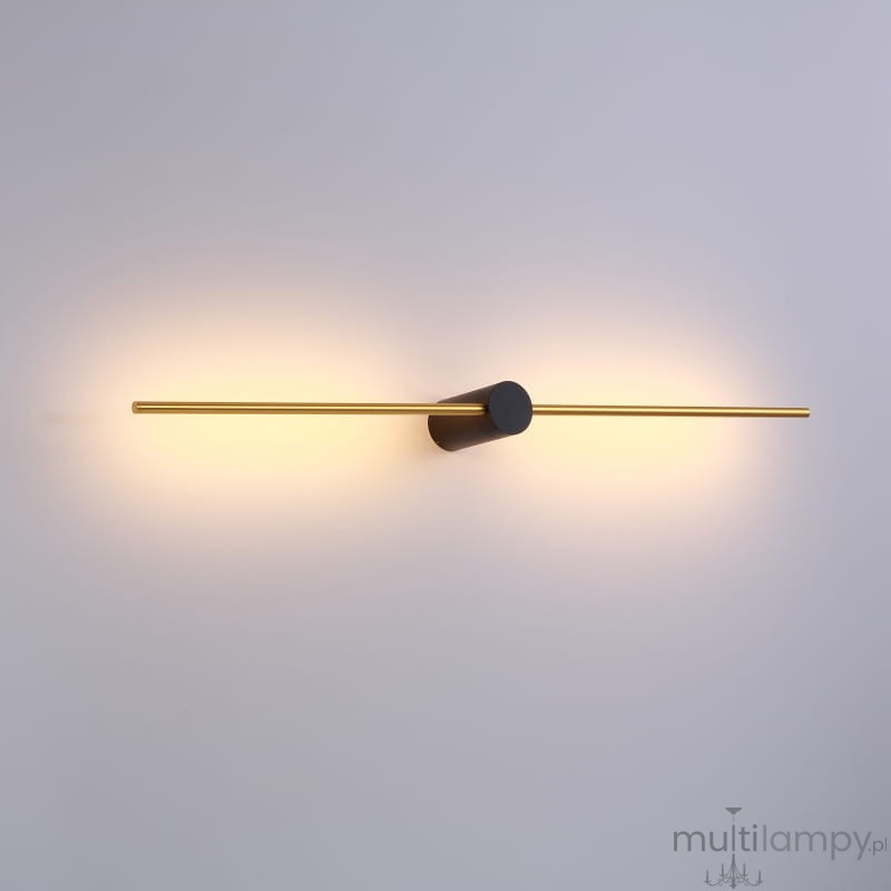 Kinkiet Wand 467 z dwoma źródłami LED 10W, w kolorze czarnym i złotym. Prosty, poziomy kształt z okrągłym mocowaniem pośrodku. Emituje ciepłe światło, montowany na ścianie, na jasnym tle.
