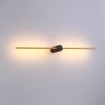 Kinkiet Wand 467 z dwoma źródłami LED 10W, w kolorze czarnym i złotym. Prosty, poziomy kształt z okrągłym mocowaniem pośrodku. Emituje ciepłe światło, montowany na ścianie, na jasnym tle.